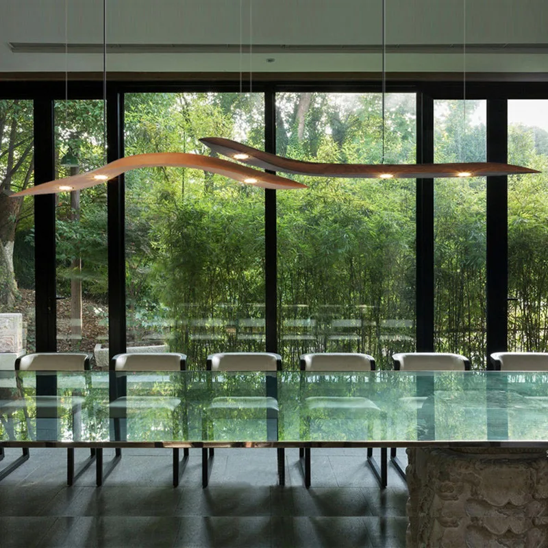 A modern dining room features a long glass table, light-colored chairs, large windows with views of greenery, and an organic touch from the Spiral Wood Pendant Light.