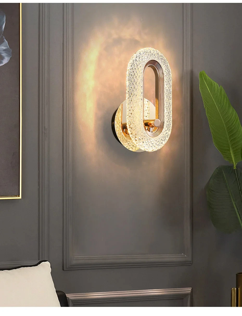 The Luxury Wall Sconce, featuring a modern and stylish oval loop design, emits a warm glow against a dark gray wall; framed artwork is partially visible on the left and a green plant sits to the right.