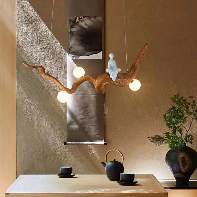 The Zen-Inspired Wooden Pendant Light, handcrafted from natural wood with round bulbs, is suspended above a table adorned with a teapot and cups. A sculpture of a seated figure on the fixture enhances the room's zen aesthetic, complemented by art and greenery.