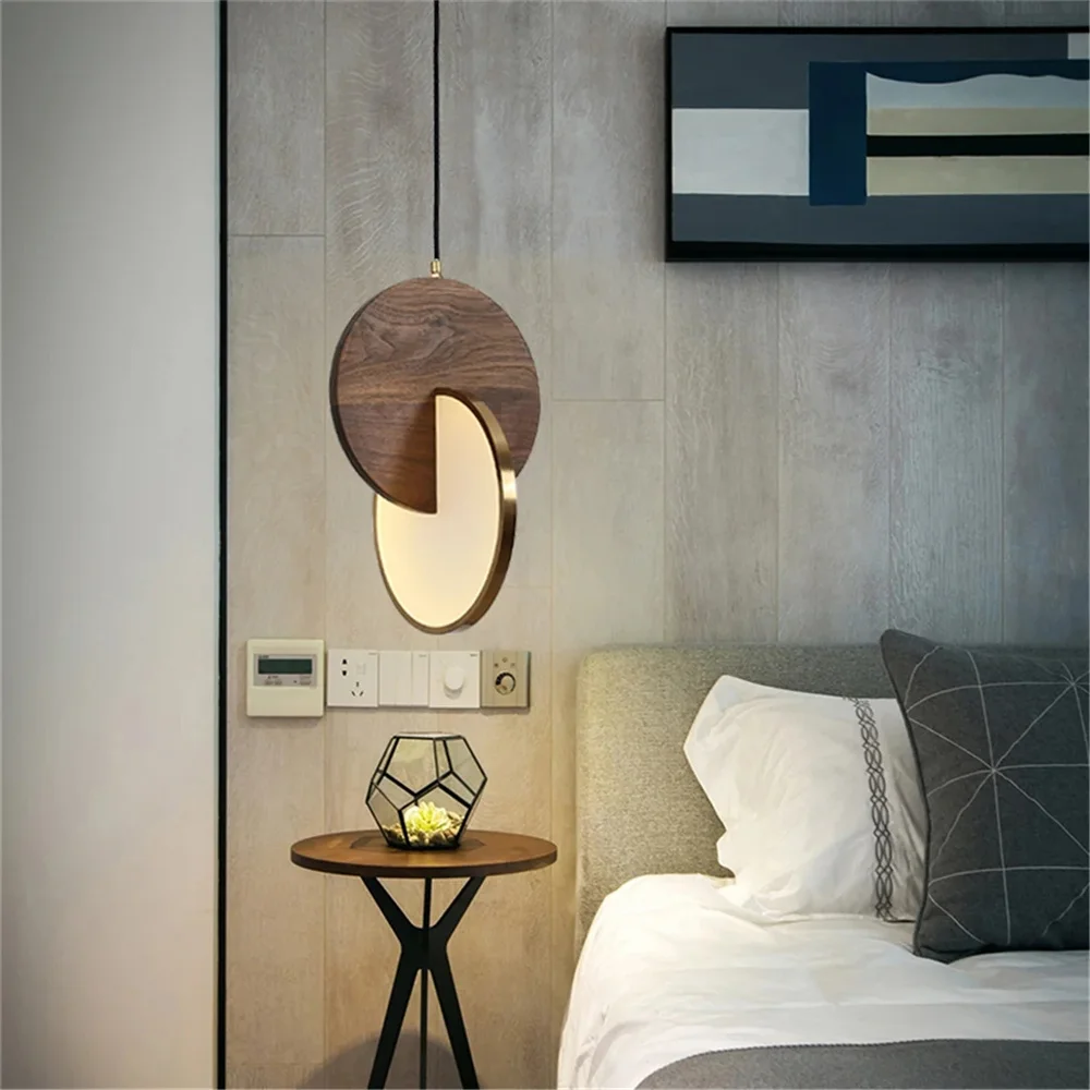 The modern bedroom exudes sophistication with a Dual Disc Pendant Light above a round side table. The table holds a geometric vase with a small plant, while gray and white bedding sets the scene on the bed. Abstract art gracefully adorns the wall.