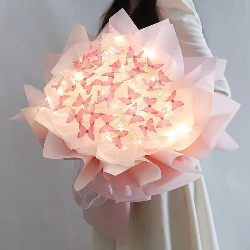 A person elegantly dressed in a white outfit holds the Creative Handmade Butterfly Bouquet, crafted from soft pink paper and adorned with vibrant pink butterflies and glowing lights. This masterpiece, part of the DIY Bride Wedding Decor Flower Material Package, resembles a flower with delicate, layered petals—perfect for home decoration or as a romantic Valentine's Day gift.