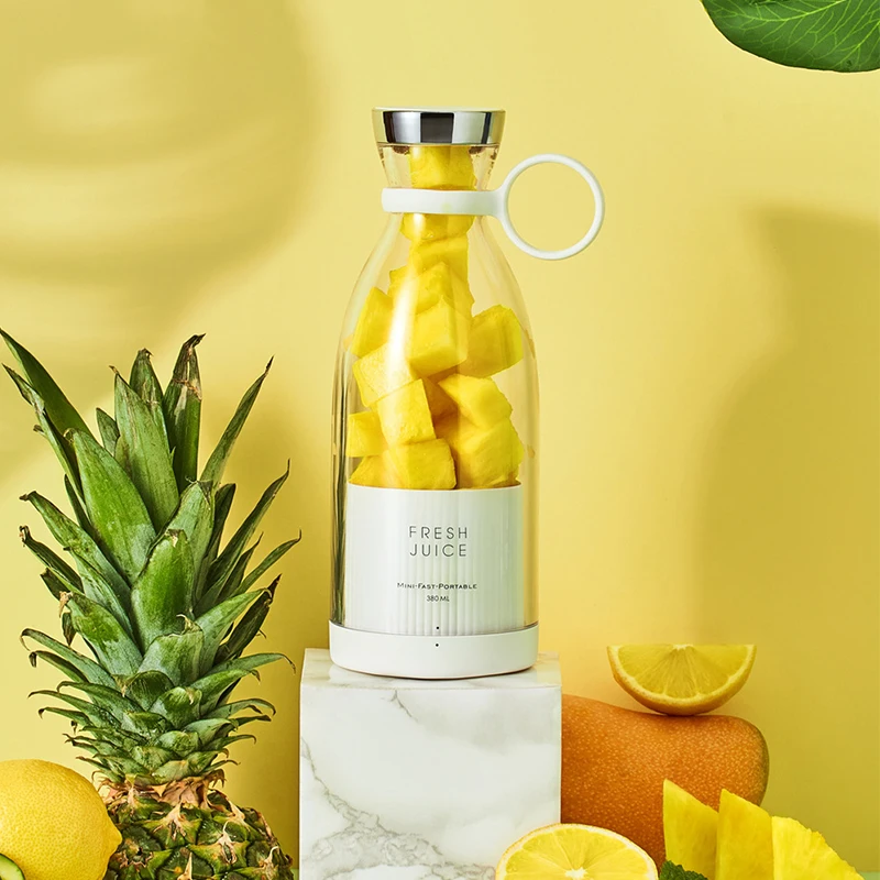 A glass bottle labeled "Fresh Juice," filled with pineapple chunks, stands on a marble pedestal, surrounded by a vibrant arrangement of pineapple, lemon, and other fruits against a yellow background. Nearby, find the convenient portable Mini Blender ready to transform those fresh ingredients into a delicious drink.