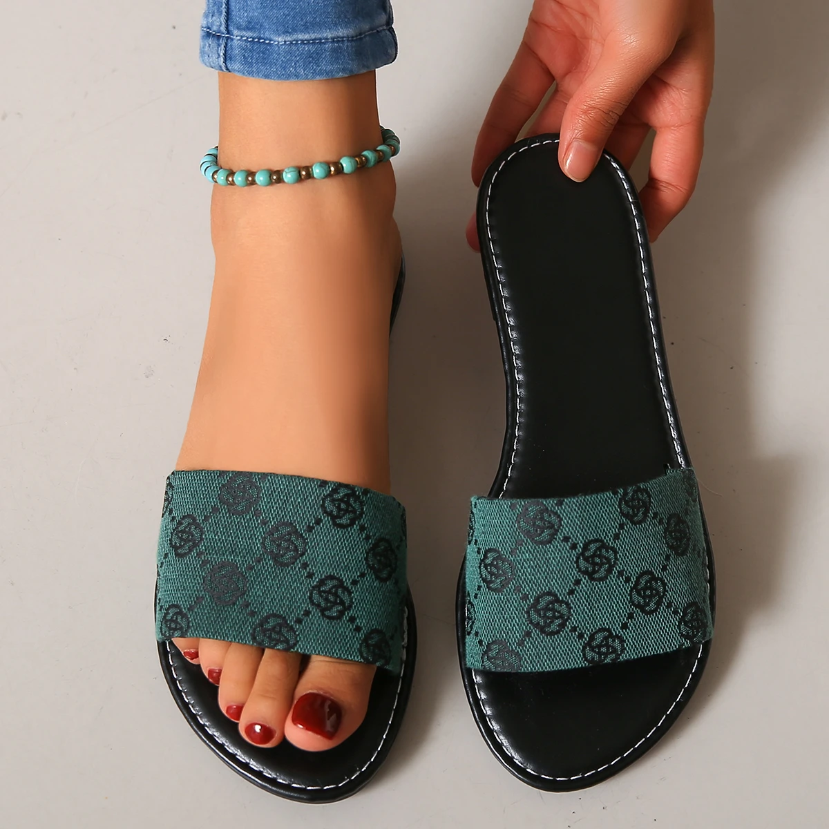 Wearing blue jeans and a turquoise anklet, someone displays one foot in a green and black patterned French Sole slipper while holding the matching sandal. Toenails are painted dark red, highlighting chic British style.