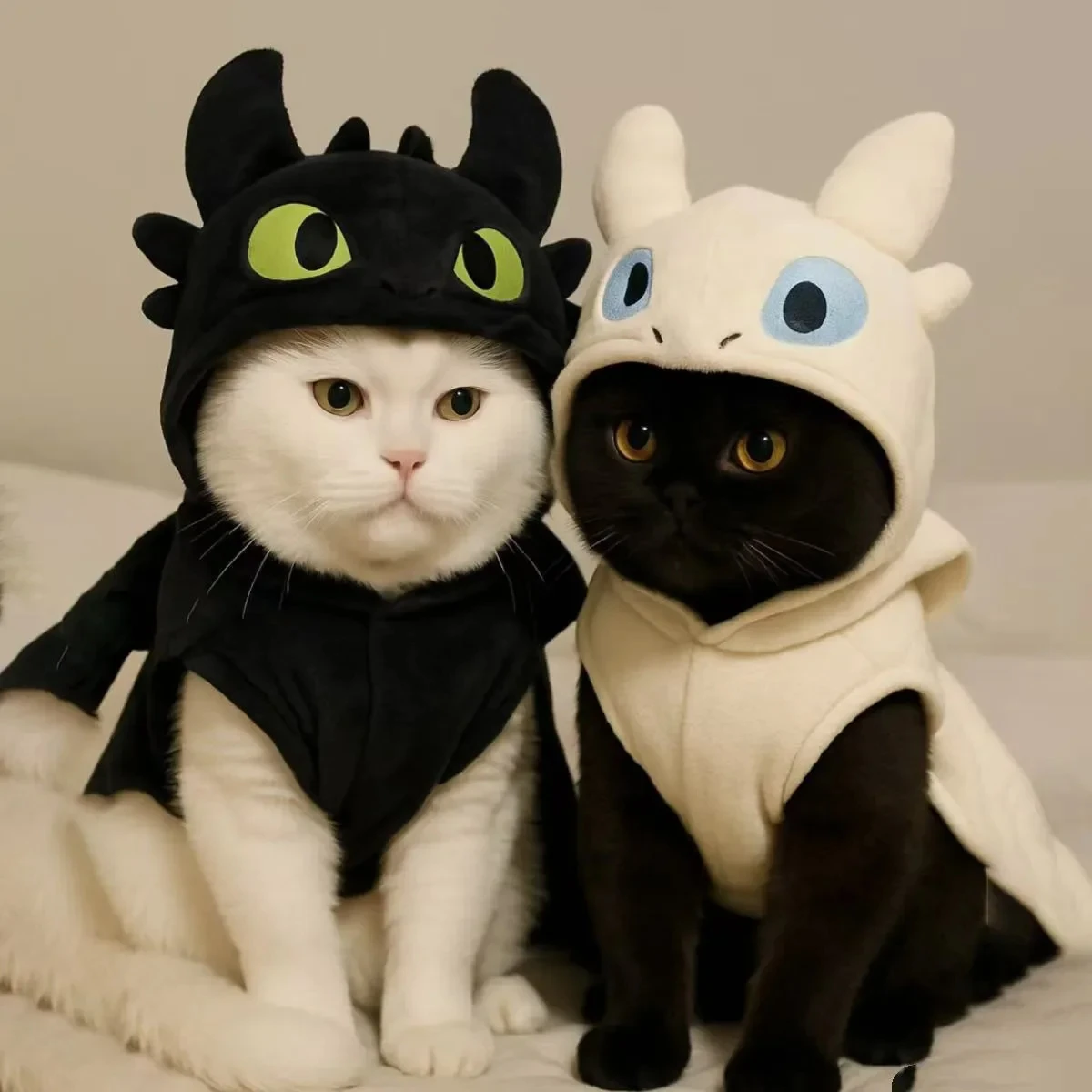 A white cat and a black cat sitting side-by-side; the white cat wears a black Pet Hoodie and the black cat wears a cream-colored Pet Hoodie, mimicking the "Night Fury" and "Light Fury" characters.
