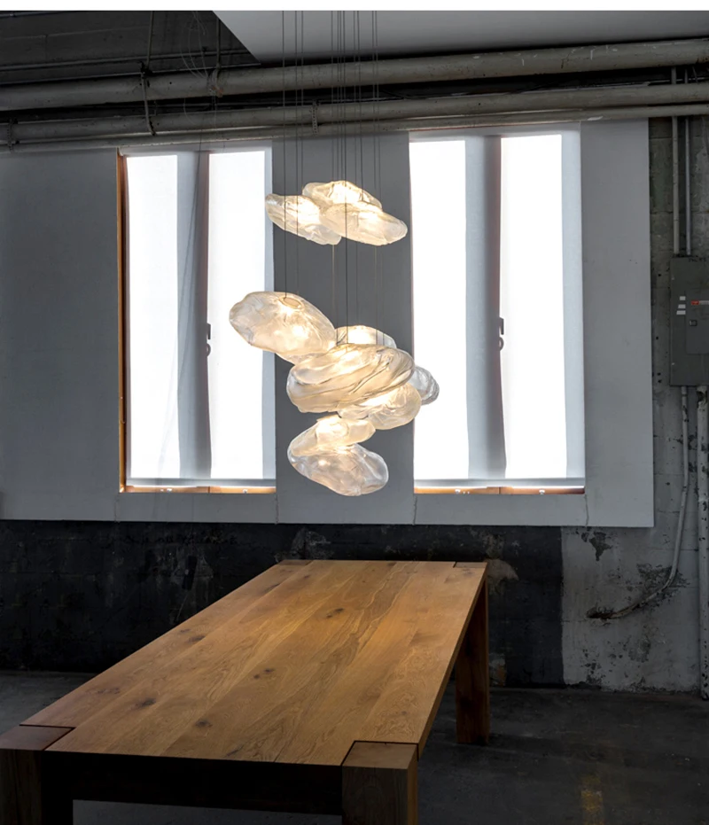 The Luxe Cloud Glass Pendant Lights float above a wooden table in an industrial-style room with tall windows and concrete walls.