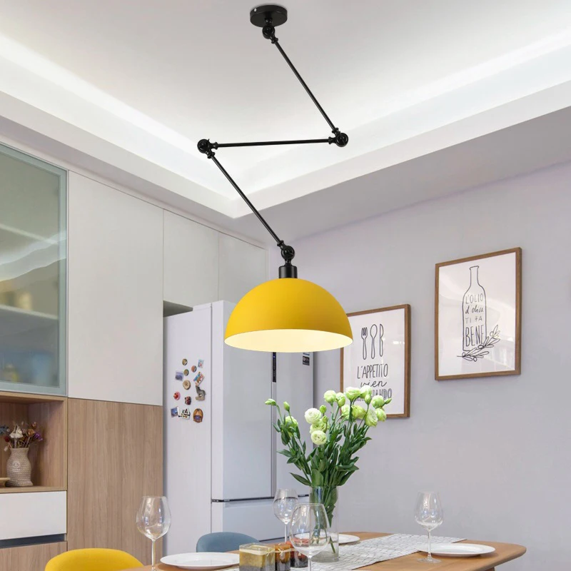 An industrial-style dining area showcases a round wooden table for four beneath a Zig Zag Pendant Light. The walls are adorned with art, and in the background, white cabinets and a refrigerator complete the modern look.