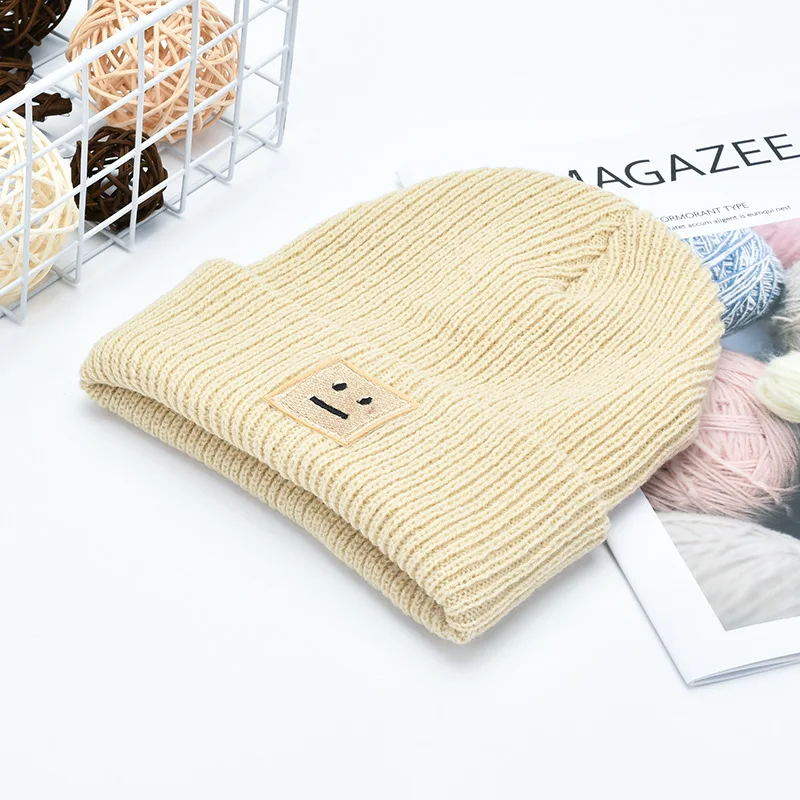 A soft beige ribbed knit men’s winter beanie hat featuring a small embroidered smiley face patch.