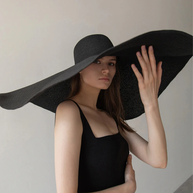 grand chapeau surdimensionné à large bord pour femmes noir, porté par une femme avec robe noire sur fond gris .Une main qui soutient le chapeau.