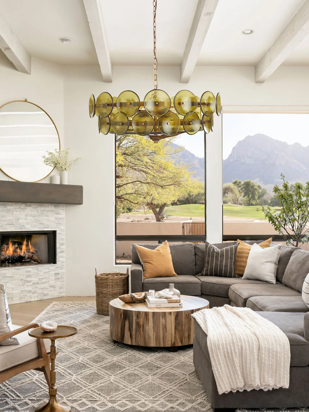 A modern living room with a gray sectional sofa, round wooden coffee table, fireplace, large windows, and an Italian Retro Pendant Light chandelier, featuring mountain views outside.