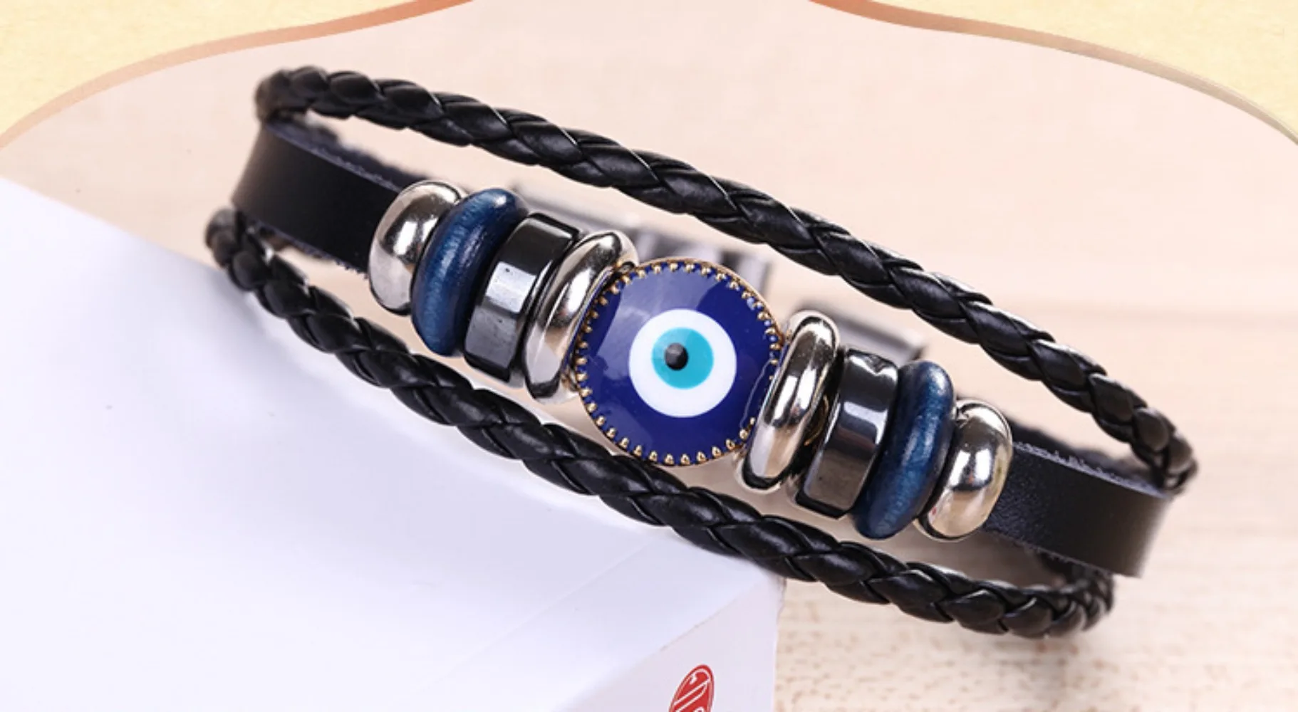 Black braided leather bracelet with blue evil eye charm on a beige background