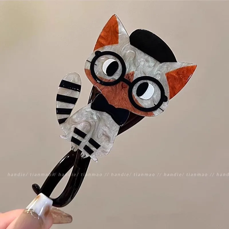 A hand holds The Acrylic Cat Hairpin from KittyNook Cat Co., shaped like a cartoon cat with glasses and bow tie.