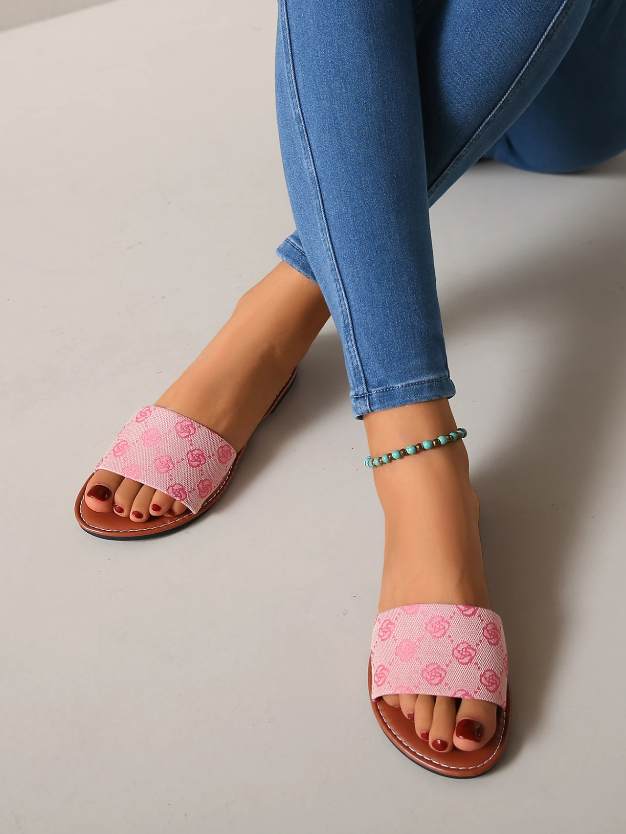 A person sits cross-legged in blue skinny jeans and pink French Sole slippers with a patterned design—summer-ready chic. Red nail polish and a turquoise beaded anklet complete this charming British-inspired look.