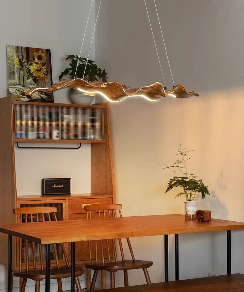 A wooden dining table with four chairs is lit by the Leaf Pendant Light. In the background, a hutch displays a vintage radio and a painting, while a plant decorates the table.