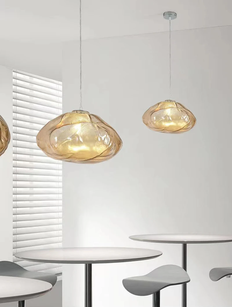 Two Luxe Cloud Glass Pendant Lights with translucent, amber-colored shades hang above round white tables and gray stools in a modern, minimalist room with white walls and a window with blinds.
