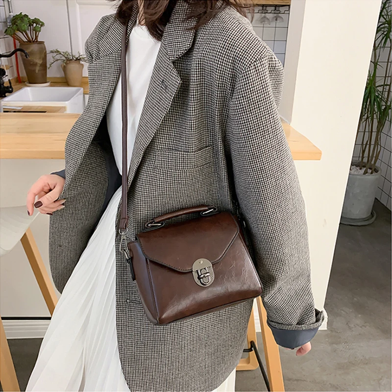 Stiilne naine kannab õlgadel väikest pruuni nahast crossbody kotti sangaga, neutraalsetes toonides ruudulise bleiseri ja valge kleidi taustal.