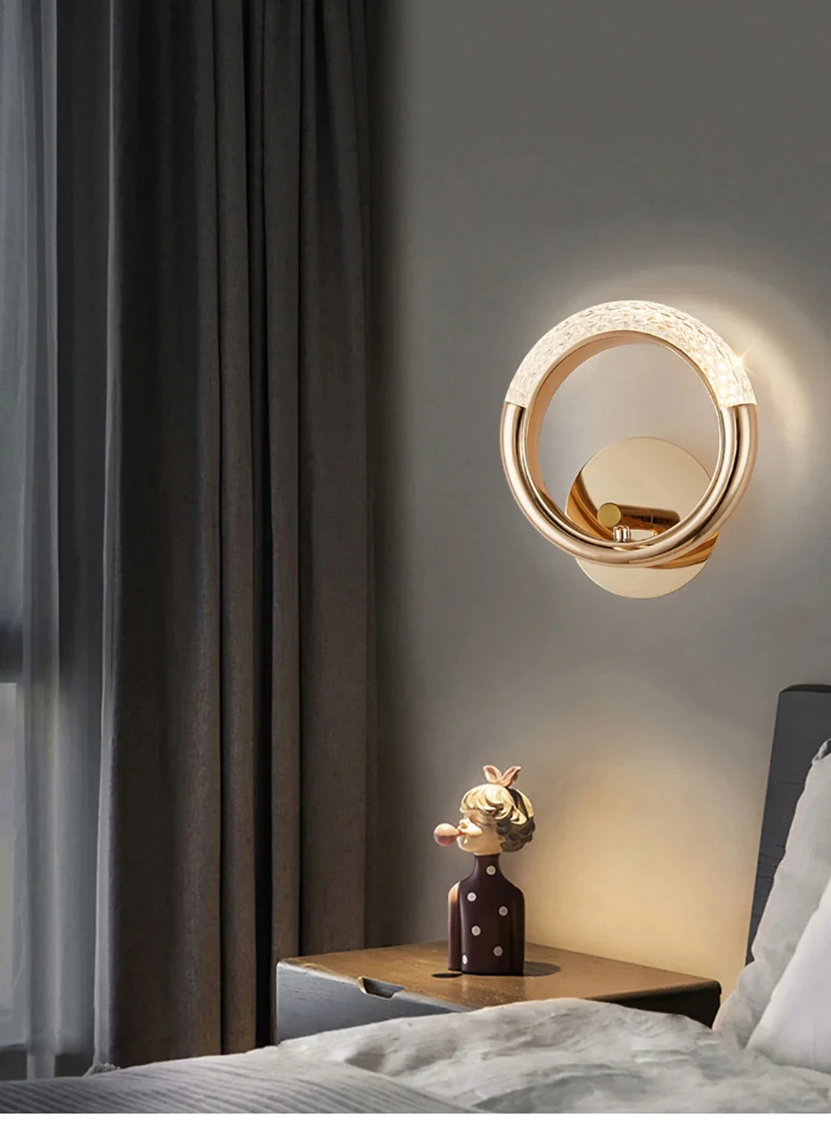 A Luxury Wall Sconce, modern and stylish with a gold circular design and frosted section, is mounted above a bedside table. The table showcases a decorative figurine adorned with a bow and polka dot outfit. Curtains and part of the bed are visible, enhancing the chic indoor lighting ambiance.
