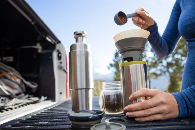 Close-up of a person pouring ground coffee into a portable coffee filter with coffee gear on an outdoor vehicle tray.
