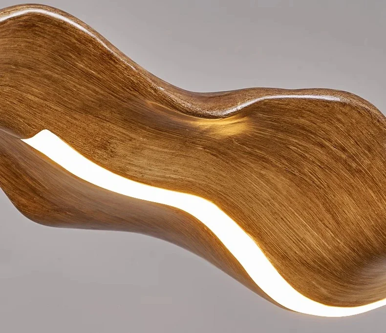 The Drift Wood Pendant Light is a wooden abstract sculpture with a wavy form and integrated light strip, enhancing its textured surface. It stands gracefully against a plain background, exuding the charm of organic-inspired design.