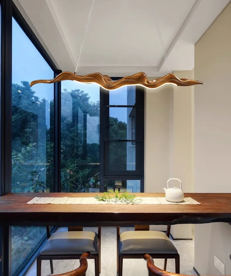 A modern dining area features a wooden table, two chairs, a white teapot, and the Leaf Pendant Light mirroring a wavy wooden fixture. Large windows display greenery outside.