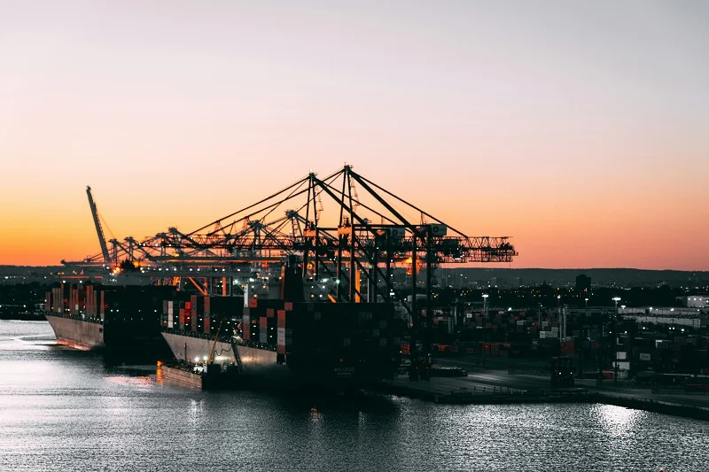 Cargo ships and cranes at dusk in a busy harbor.