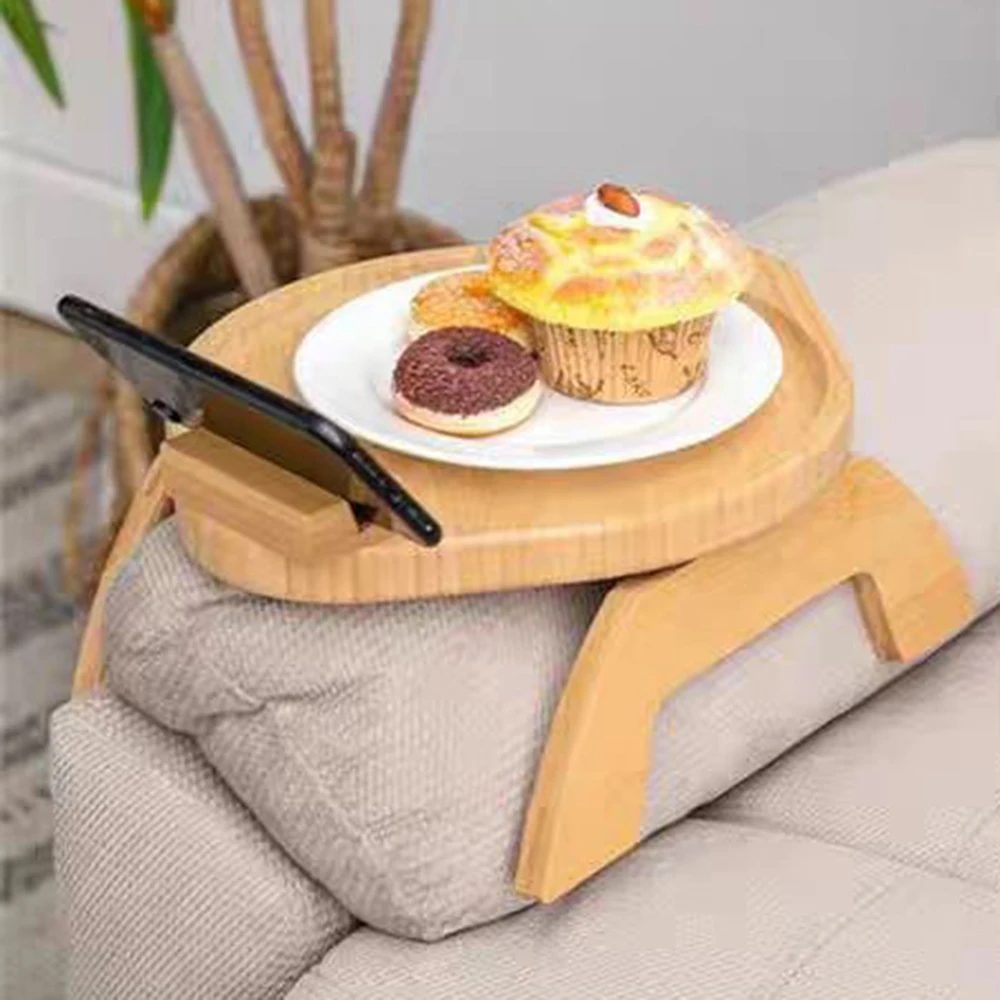 A Tray for Sofa, stylishly crafted from bamboo and wood, sits elegantly on the couch supporting a plate with a muffin and doughnut. Designed for convenience, it features a slot perfect for holding a smartphone while a plant adds charm in the background.