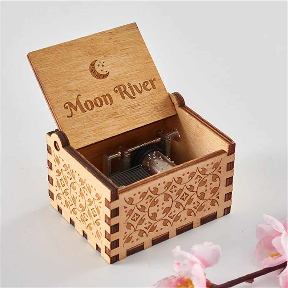 A hand-cranked Wooden Music Box adorned with intricate carvings and "Moon River" inscribed on the lid. The lid, when opened, reveals the music mechanism inside that plays classic tunes. With pink flowers in the foreground accentuating its charm, it stands out as one of the finest handmade music boxes.