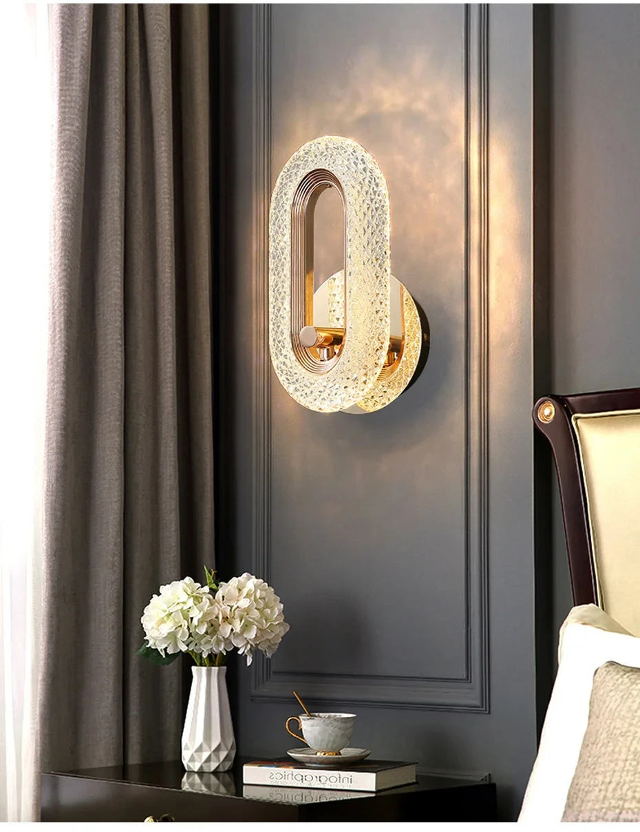 A modern and stylish Luxury Wall Sconce with an oval design is mounted on a dark wall above a bedside table, which holds a white vase with flowers and a small plate. Part of a bed with a light headboard is visible, showcasing the sleek indoor lighting solution.