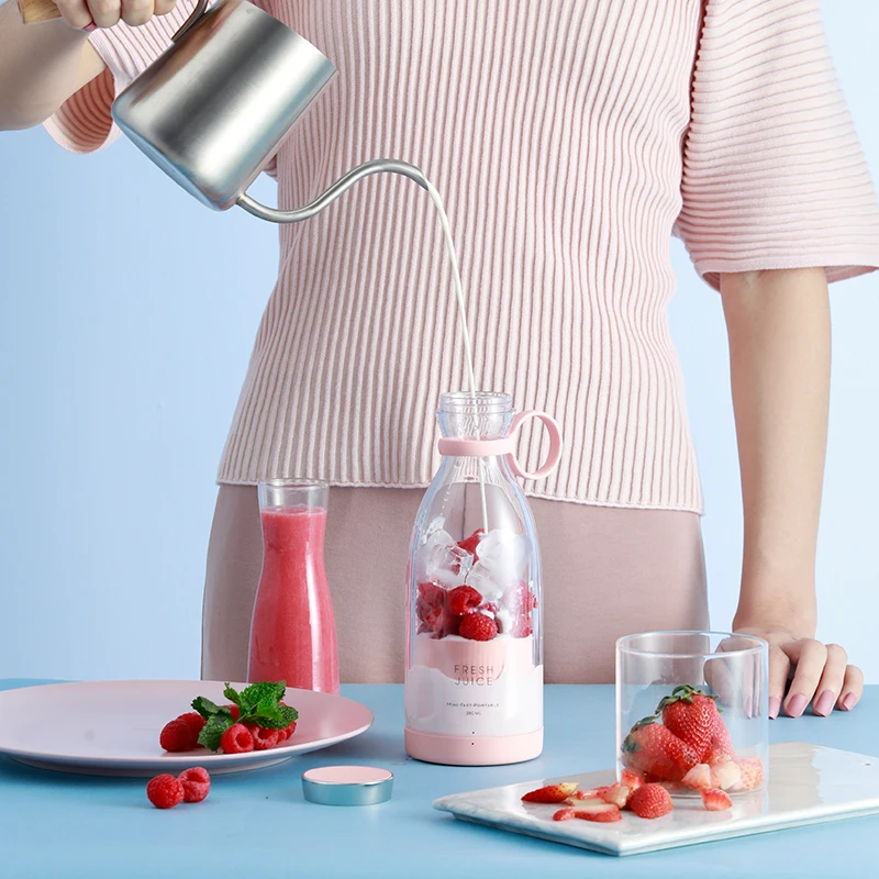 A person wearing a pink sweater is pouring liquid from a silver watering can into a bottle filled with strawberries and ice. On the table, there is a glass, a plate of strawberries, and a jar of pink liquid, all placed next to the Mini Blender, which is ready for action.