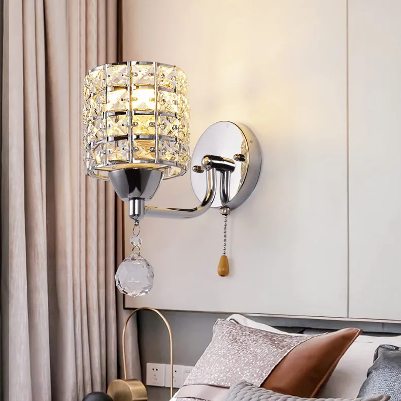 The Luxury Cylinder Wall Light with a modern shade and hanging crystals is mounted above a bed with decorative pillows, bringing luxurious lighting to this contemporary bedroom.