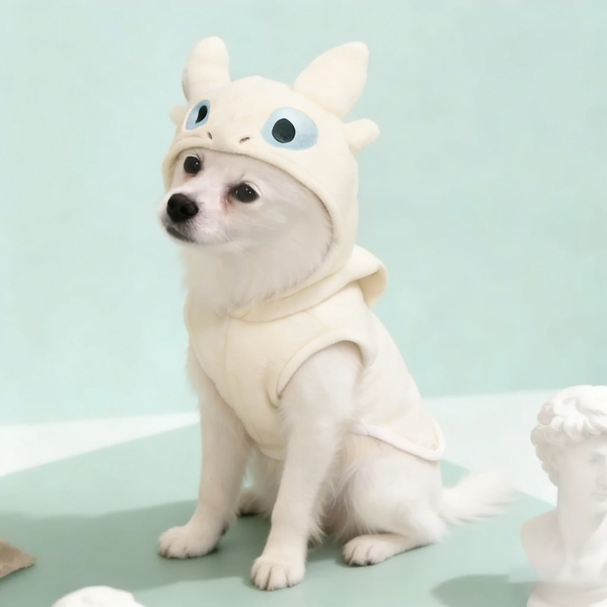 A small white dog sitting on a light blue surface next to white statues, wearing a cream-colored dragon Pet Hoodie with soft blue accents.