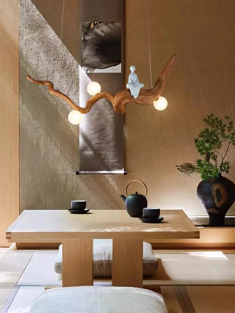 A minimalist tea room features a wooden table, two cushions, and a Zen-Inspired Wooden Pendant Light casting a soft glow. A plant and teapot on the table complete the serene setting, embodying organic beauty.