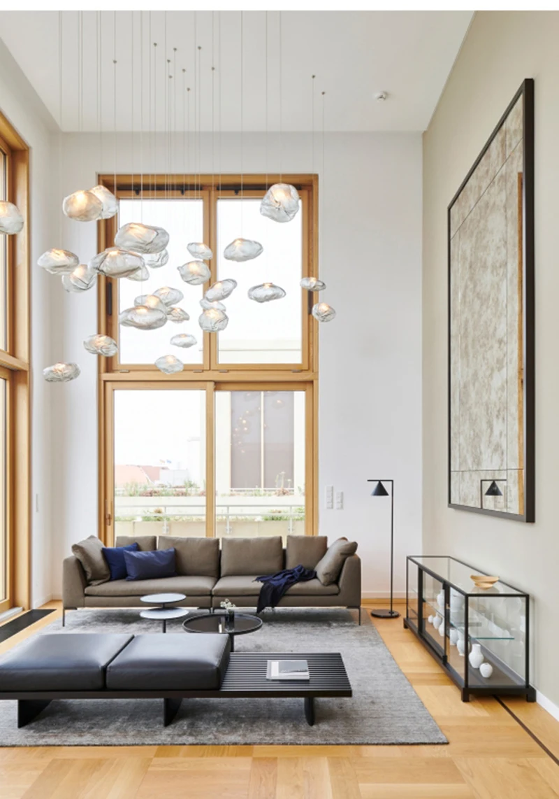 Modern living room with large windows, sofa, glass display cases, and coffee tables. The Luxe Cloud Glass Pendant Light and a large wall art piece add character. Light wood flooring and neutral tones prevail throughout the space.