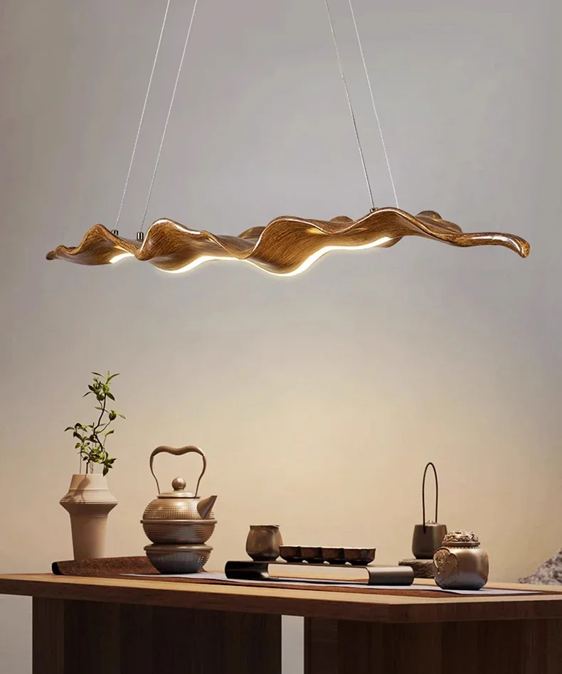 The Drift Wood Pendant Light, featuring a modern organic design with a wavy texture, hangs above a dining table adorned with teapots, cups, and decorative items against a neutral backdrop.